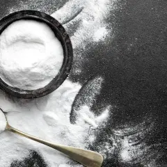 Biały proszek (soda oczyszczona) w miseczce i na łyżce na czarnym tle.