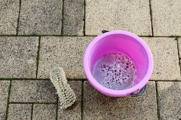 Różowe wiaderko z pianą i szczotka na kostce brukowej. Czas na czyszczenie kostki brukowej octem, by pozbyć się mchu.